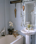 A detail of traditional bathroom with a large silver framed mirror above a sink and next to a bath with a gold taps and shower controls