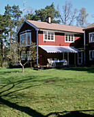 Außenansicht der Rückseite eines traditionellen Holzhauses mit einer Markise über einem Tisch und Stühlen mit Blick auf eine große Rasenfläche