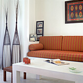 Stone built sofa with striped chenille cushions in a white living room with pinewood table and rattan artefacts