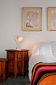 Lampe auf geschnitztem Nachttisch neben Bett mit gestreifter Bettdecke und gerahmten Kartografiken