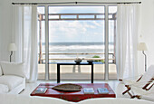 Wohnzimmer mit großen Fenstern mit Blick auf das Meer mit Leinenvorhängen und einem rot lackierten chinesischen Holztisch mit goldenen Motiven