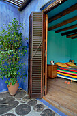 Aquamarine bedroom with beamed ceiling and Mexican blanket on bed viewed through shuttered doorway