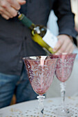 Mann öffnet Weißwein und zwei verschnörkelte rosa Weingläser