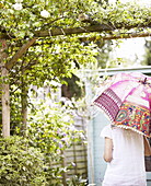 Frau im Schatten eines bunten Sonnenschirms unter einer Pergola mit Kletterrosen in einem Londoner Garten UK