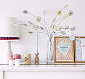 Paper flowers and framed book covers on mantlepiece shelf with lamp in Bussum home, Netherlands