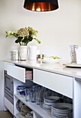 Glasses and plates on shelf in kitchen unit with cut flowers and black pendant lamp, Oxfordshire, England, UK