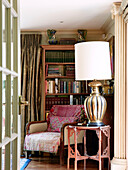 Large brass lamp on side table with vintage armchair and bookcase in traditional country house Welsh borders UK