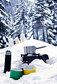 Kochtopf auf Gaskochfeld mit Flachmann und Tassen im Schnee, St. Anton, Tirol, Österreich