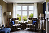 Blue and brown checked armchairs with lamps in window of Northumbrian coastal home, UK