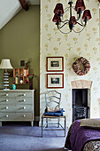 Red lamps in Cotswolds bedroom with floral patterned wallpaper and upcycled chest of drawers and chair, UK