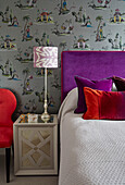 Purple headboard and Oriental wallpaper with red velvet pillows and chair in London apartment, UK