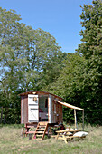 A Shepherds Hut in a summer meadow with a canvas canopy for a makeshift kitchen diner and open door with steps to the room inside