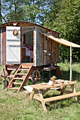 A Shepherds Hut in a summer meadow with a canvas canopy for a makeshift kitchen diner and open door with steps to the room inside