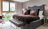 Double bed with buttoned headboard and footstool beside picture window in Lakes home, England, UK