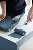 Woman roller painting sideboard top with blue paint in UK home