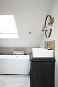 Runder Spiegel über Waschbecken mit freistehender Badewanne unter Dachfenster in Dachgeschoss-Badezimmer in Brighton, East Sussex, UK