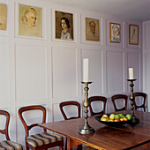 Esszimmer mit Esstisch und Stühlen aus Eichenholz, Kerzenleuchtern aus Zinn und einer Obstschale mit Äpfeln, lila bemalten Holzpaneelen und Porträts an der Wand