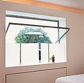 Offenes Fenster mit Metallrahmen in einem modernen, weiß getäfelten Schlafzimmer mit mattiertem Plexiglasfenster und Kaktus