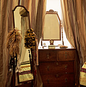 Schlafzimmerdetail mit Chevalspiegel und Federschmuck neben antiker Kommode mit ungeschnittenen Seidenbandvorhängen