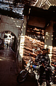 Man sitting on his motorbike in dappled sunlight in a covered alleyway in the Medina Fez Morocco