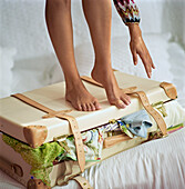 Young woman standing on an over packed suitcase on a bed