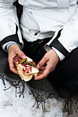 Frau in Skijacke sitzt und isst Salami und Gewürzgurken auf Brot in Zermatt, Wallis, Schweiz