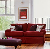Red sofa with purple cushions and footstool under modern artwork between sash window and open door to balcony