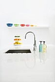 Kitchen sink with chrome mixer tap built into white worktop with bottles of soap a shelf of colourful bowls and a cakestand with citrus fruit