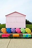Pink beach hut on grass bank with multi coloured umbrellas in a row and beach towels