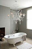Rolltop-Badewanne und überdimensionaler neobarocker Kronleuchter mit Kerzenlicht in einem grau gestrichenen Badezimmer mit Steinbodenfliesen