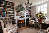 Armchairs at lit fireside with bookshelves and Christmas tree in London home  England  UK
