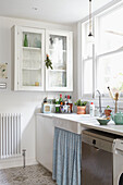 Glass fronted cabinet above sink with curtain at kitchen window in London home  UK