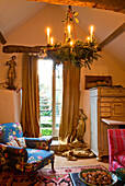 Candle lit chandelier decorated with holly and spruce hanging in a living room with open French door with gold curtains and figurines