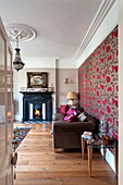 Gemusterte Tapete über braunem Sofa mit beleuchtetem Kamin im Wohnzimmer eines Hauses in Middlesex, London, England, UK