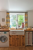 Butler sink and wooden cupboard with washing machine below kitchen window of Cambridge cottage England UK