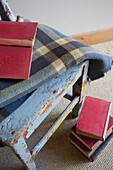 Rote, gebundene Bücher mit Schottenkaro-Decke auf verwittertem Stuhl in einem Bauernhaus in Kent, England