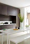 White dining table and dark wood fitted kitchen in contemporary London apartment, England, UK