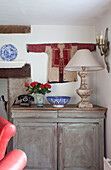 Wall mounted embroidered to with cut flowers and lamp on sideboard in Sussex farmhouse, UK