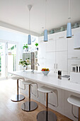 Barhocker an der Frühstücksbar in einer weißen Einbauküche mit hellblauen Hängeleuchten in einem modernen Haus in London, UK
