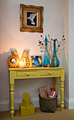 Coloured glassware and lit candle on yellow painted side table in London home, England, UK