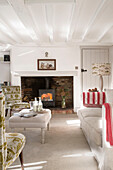 Pair of upholstered chairs and ottoman footstool with lit woodburner in beamed living room of London home,  England,  UK