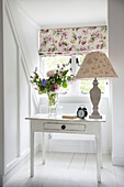 Lamp and flowers on table at window with floral blind in Sussex country house England UK