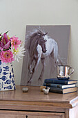 Equestrian artwork with hardbacked books and cut flowers on wooden chest in Berkshire bedroom England UK