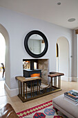 Schwarzer runder Spiegelrahmen über einer offenen Feuerstelle mit Holzofen in einem Haus in Gloucestershire, England UK