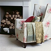 Floral print loose cover on an armchair in front of a fireplace with log pile