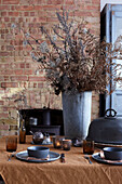 Set table with dried flowers in metal vase in front of brick wall