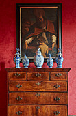Oil painting hung on a wall with red wallpaper, underneath antique chest of drawers with fine china being displayed