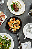 King prawns in a pan with garlic and parsley, leaf alad and a lamb chop. A group of dishes