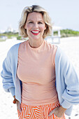 A blonde woman wearing an apricot top, a light-blue cardigan and summer trousers on a beach