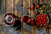 Rose hip berries herbal tea in glass teapot and cup on old wooden plank background with wild autumn berries around